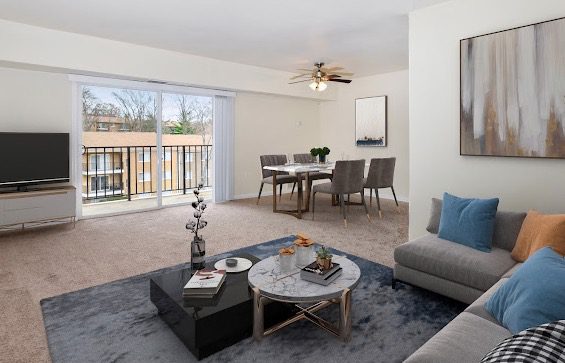A modern, open-plan living room with a sliding glass door leading to a balcony. It features a gray sofa with blue and orange cushions, a marble coffee table, a wall-mounted TV, and a dining area with a table and chairs. Neutral colors and abstract art adorn the space.